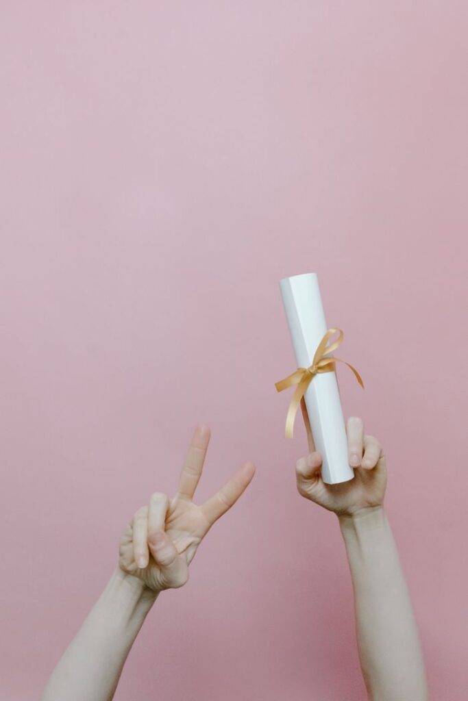 Diploma held in hand with peace sign on pastel pink background.